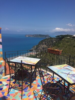 A terrace with two tables and chairs, a colorful tiled floor, and a view of the sea and a distant island.