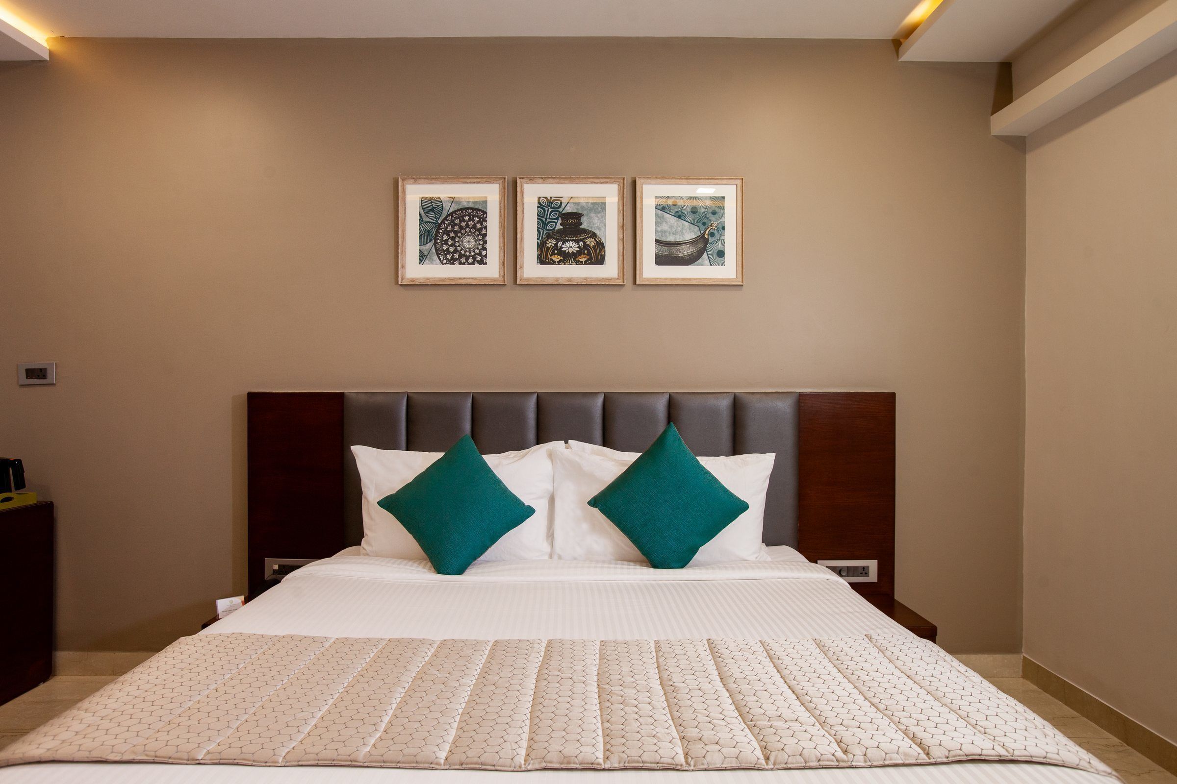 A hotel room with a large bed, two green pillows, a headboard, and three framed artworks on the wall.