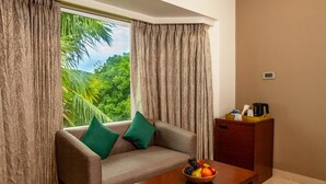 A room with a sofa, a table with a fruit bowl, a coffee maker, and a view of greenery through curtains. - Mango Suites Viera (Hyderabad)