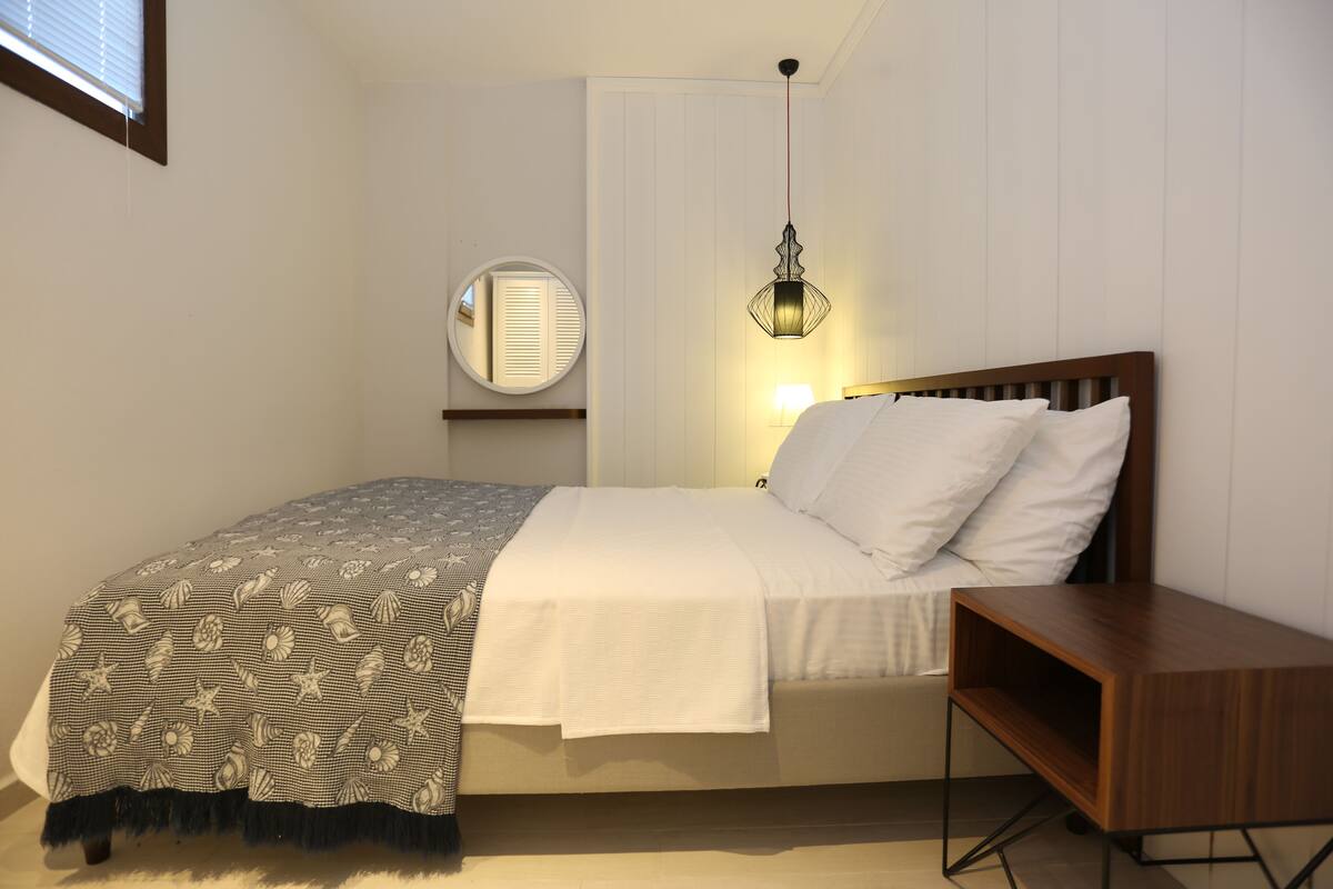 A bedroom with a bed, bedside table, lamp, mirror, and a window with blinds.