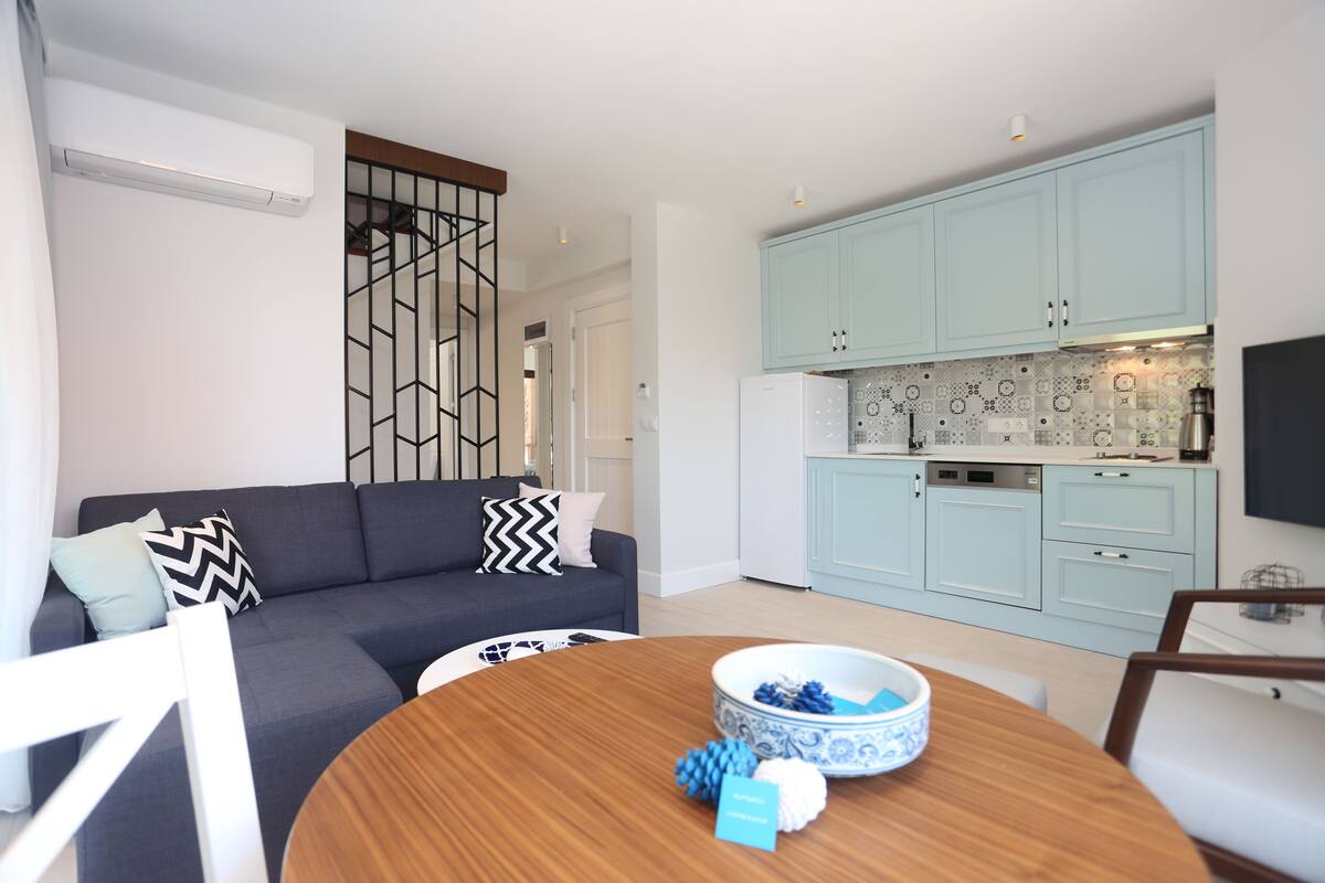 A modern living room with a dark grey sofa, a round wooden coffee table, and a kitchen area with light blue cabinets and patterned backsplash.