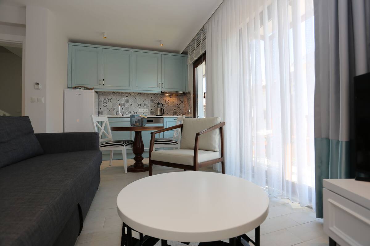 A modern living room with a grey sofa, a round white table, and a wooden chair. A kitchen with light blue cabinets and a white refrigerator is visible in the background.