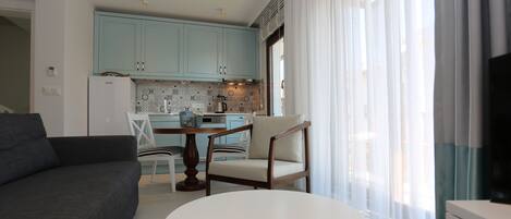 A modern living room with a grey sofa, a round white table, and a wooden chair. A kitchen with light blue cabinets and a white refrigerator is visible in the background.