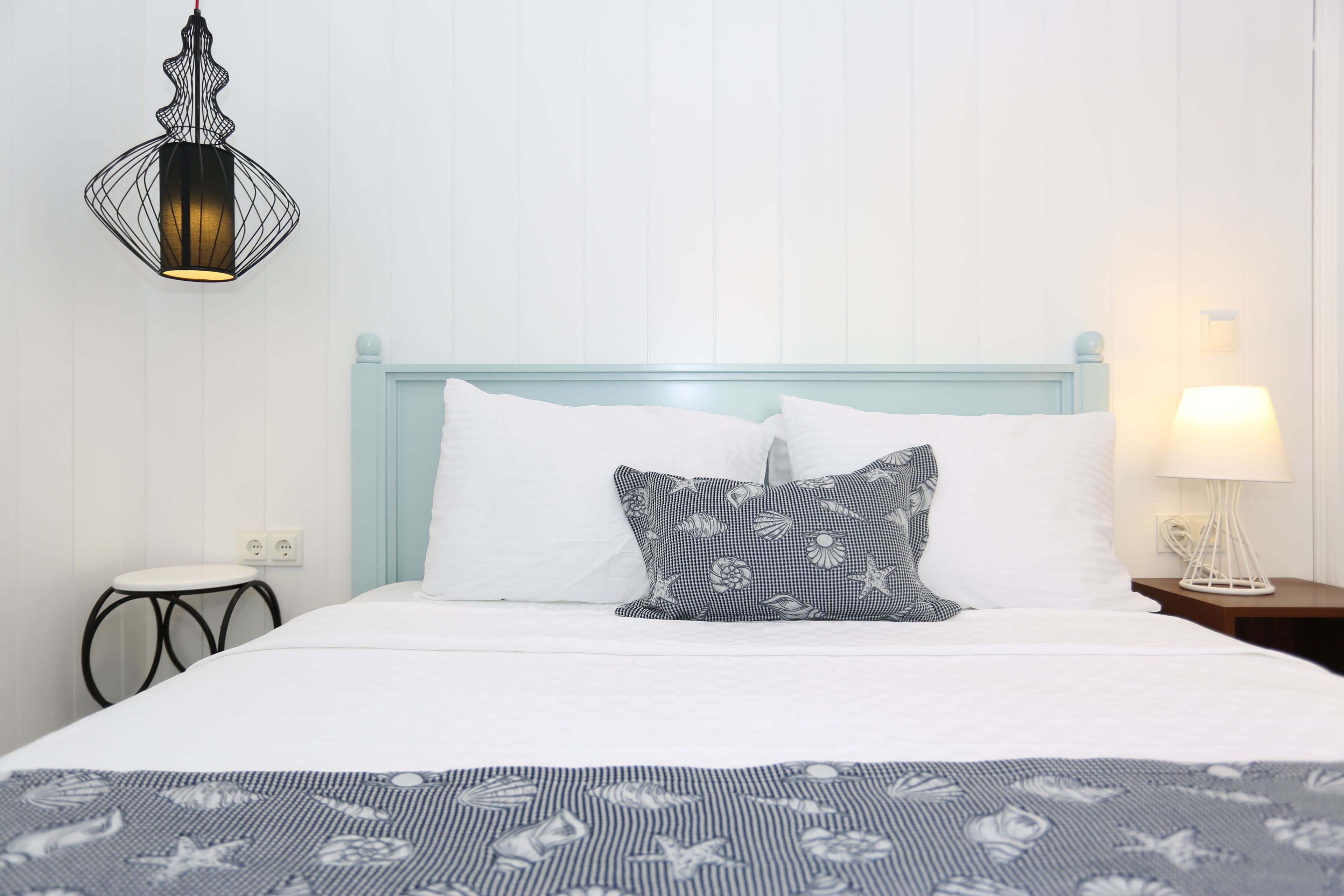 Una cama bien hecha con almohadas blancas y estampadas, una colcha azul a rayas, una lámpara de pared y una mesita de noche con una lámpara.