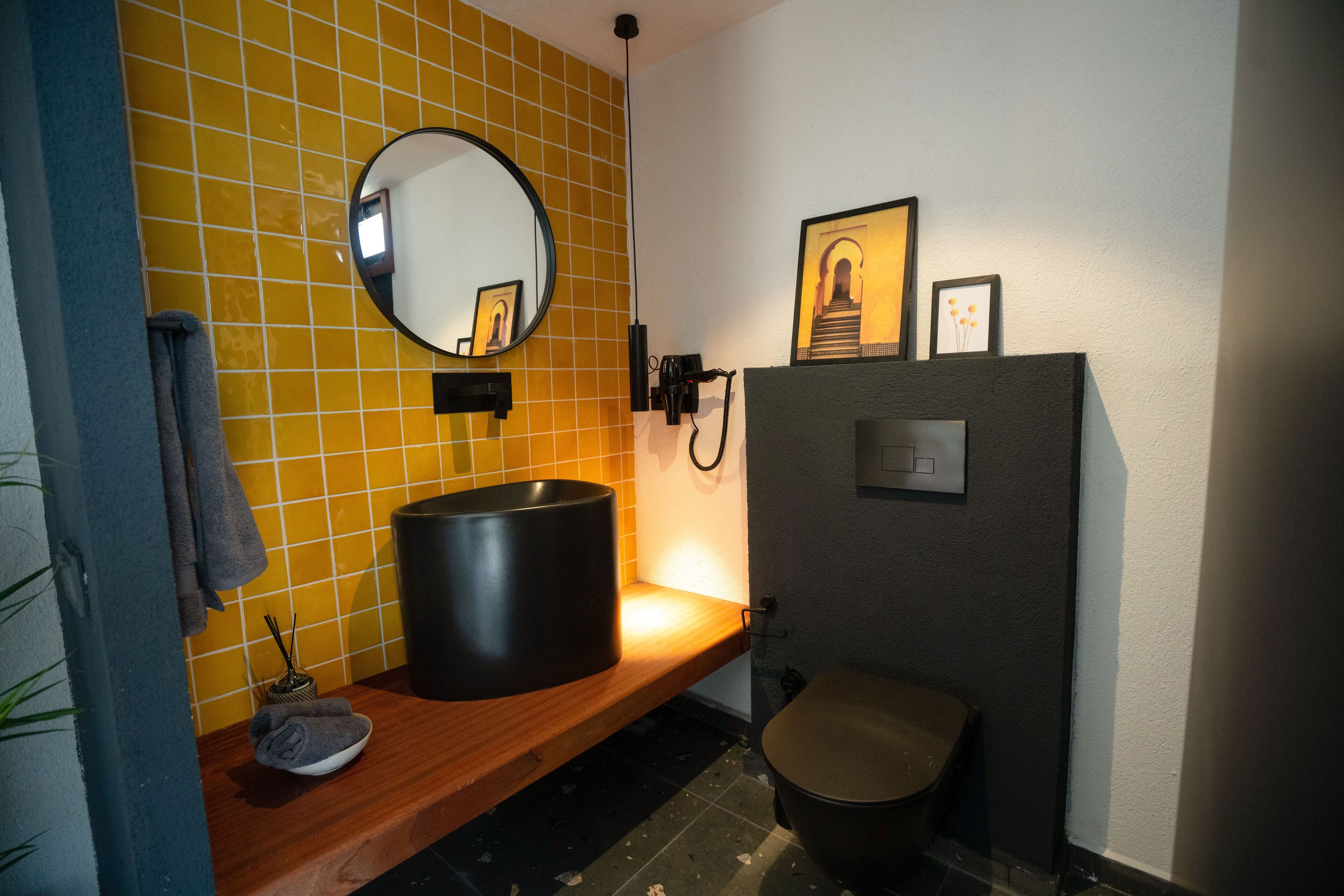 A bathroom with a yellow tiled wall, a round mirror, a wooden shelf with a bowl and a towel, a toilet, and a framed picture on the wall.