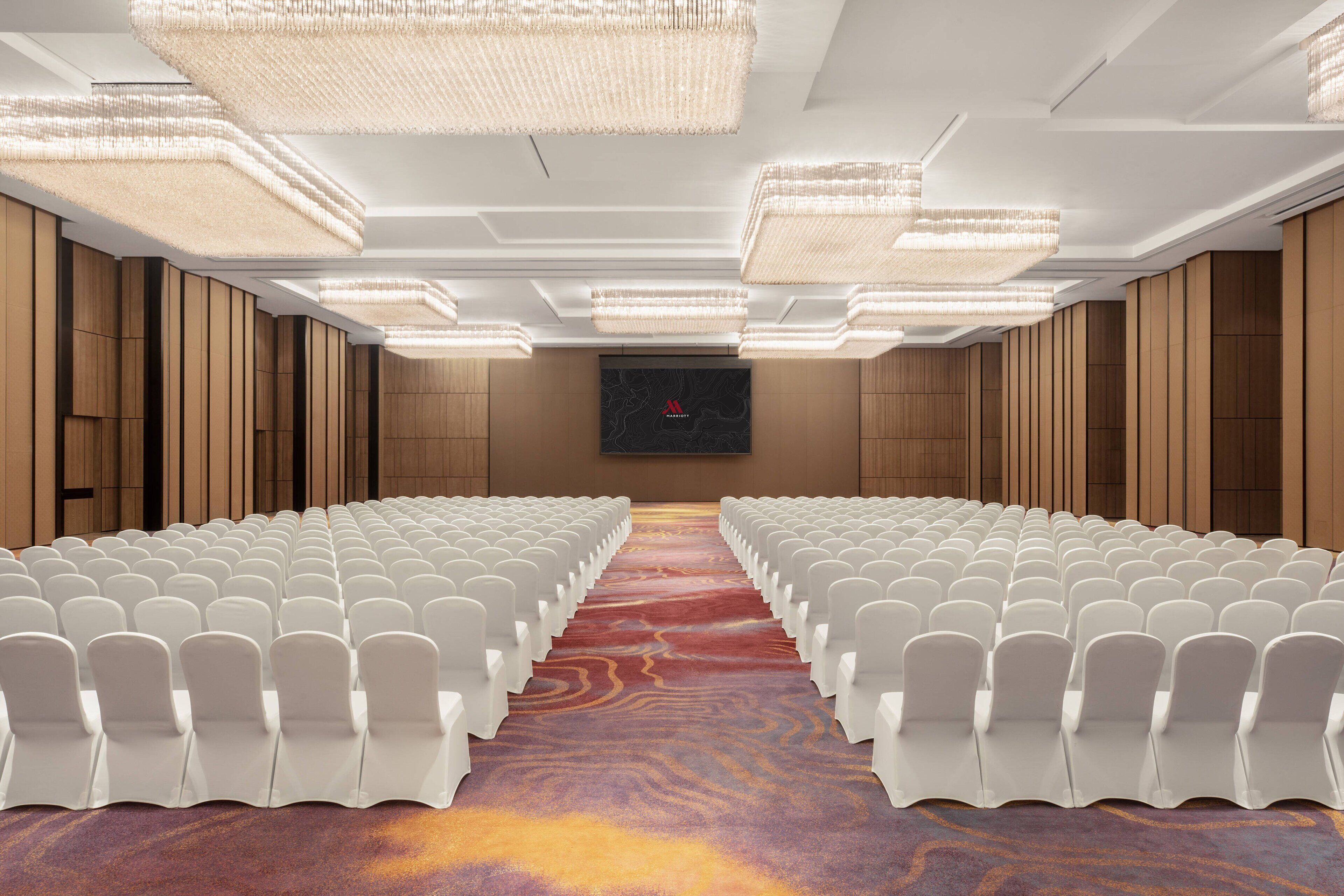 A conference room with rows of white chairs, a red carpet, and a large screen at the front.