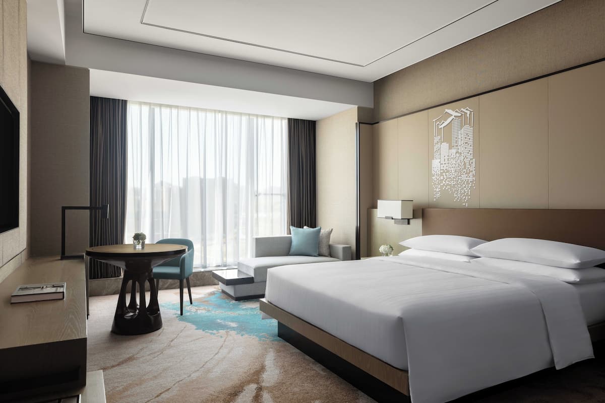 A modern hotel room with a large bed, a desk with a chair, a sofa, and a large window with curtains.