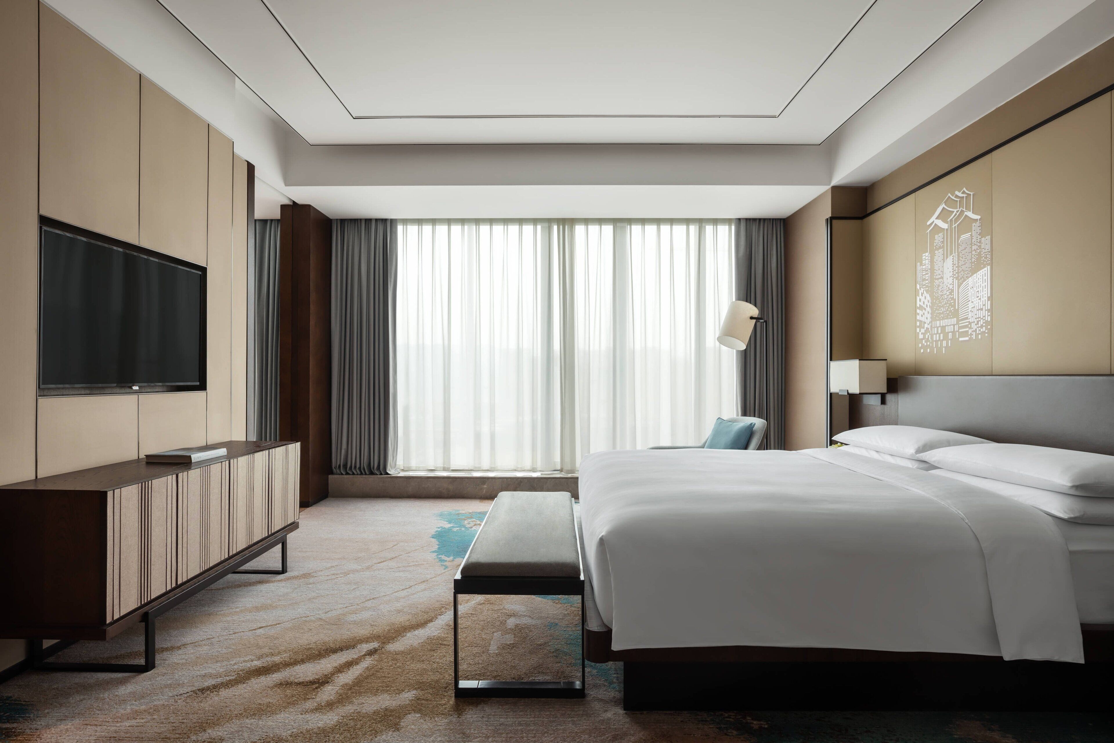 Quarto de hotel moderno com uma cama grande, televisão de tela plana, um banco e vista para a cidade através de amplas janelas.
