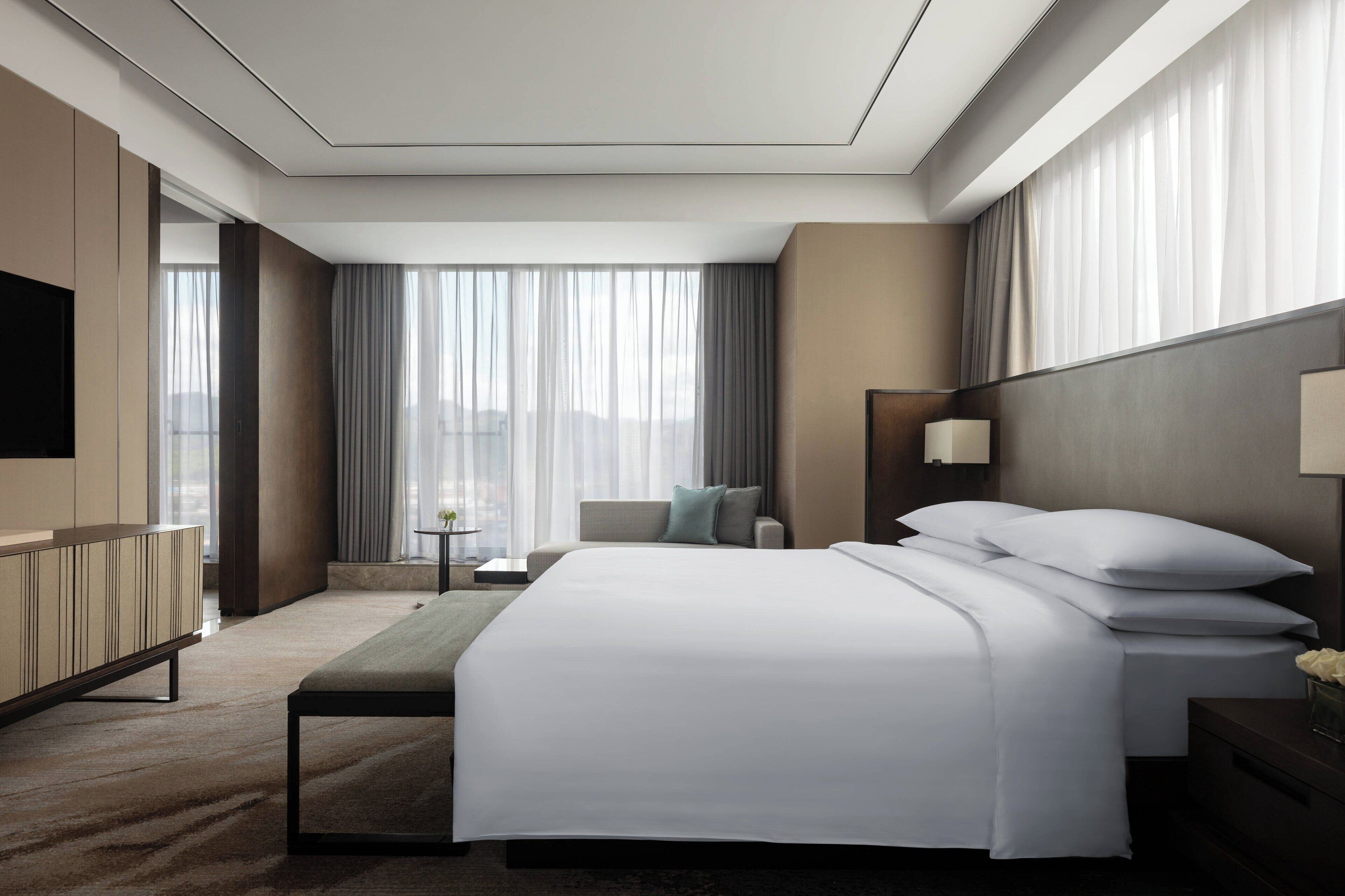 A modern hotel room with a large bed, a bedside table, a TV, and a view of the city through large windows.