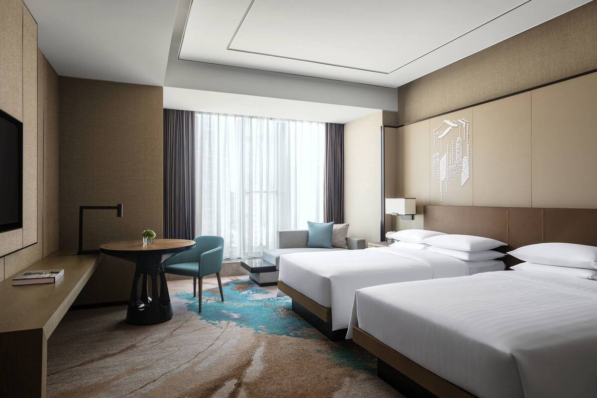 A hotel room with two beds, a desk with a chair, a TV, and a large window with curtains.