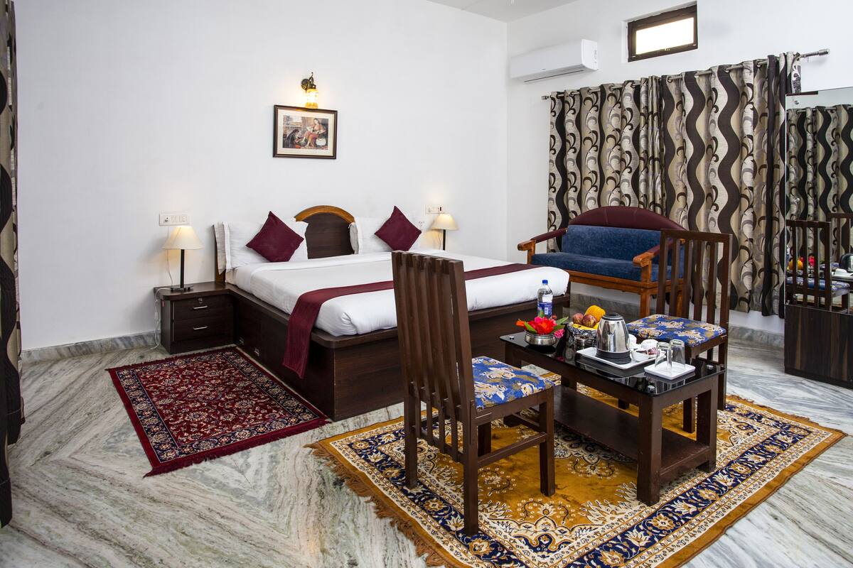 Habitación de hotel con cama, una silla de madera, una mesita con un florero y una alfombra estampada en rojo y azul.