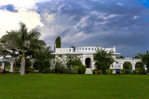 Um edifício branco com uma entrada ampla, cercado por um gramado bem cuidado e palmeiras.