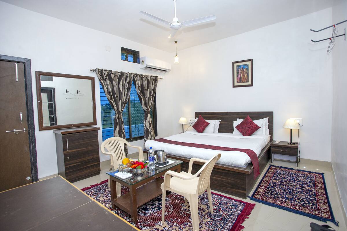 Habitación de hotel con cama, escritorio con silla, mesa con frutas y hervidor, espejo, ventana con cortinas y ventilador de techo.