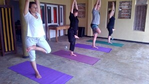 A yoga class in a room with a concrete floor and yellow walls. There are four participants on purple mats, one person standing by the wall, and a bench in the background. - Asiri Yoga Retreat (Boossa)