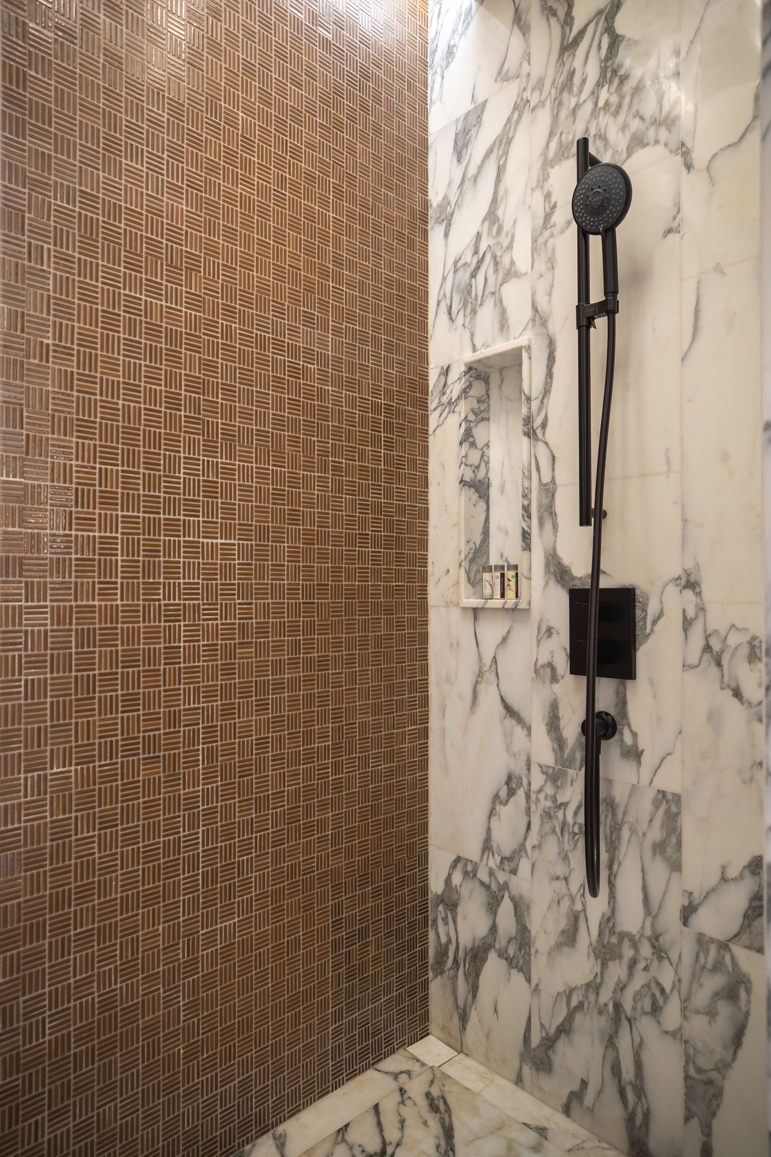 A bathroom with a patterned tile wall, a marble wall with a showerhead, and a small shelf with toiletries.