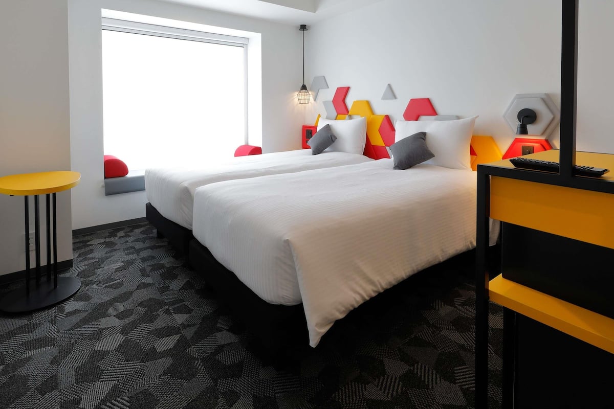 A hotel room with two beds, a desk, a chair, and a window.