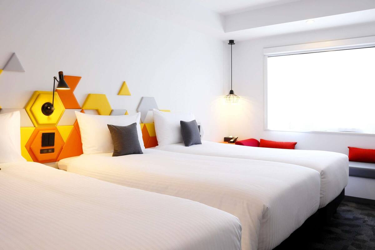 A hotel room with two beds, a geometric wall design, a window, and a lamp.