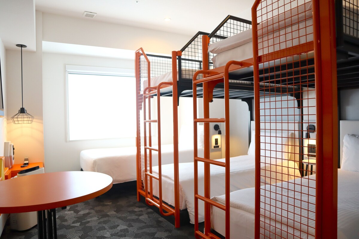 A hotel room with bunk beds, a small table, a lamp, and a window providing natural light.
