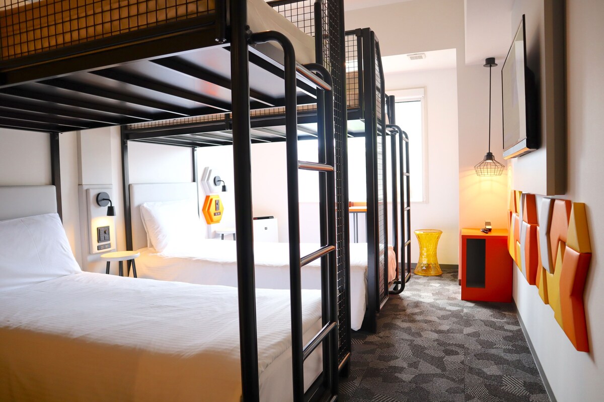 A hotel room with bunk beds, a desk, a chair, a lamp, and a wall-mounted TV.