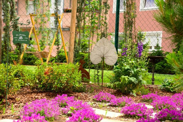 Un jardin avec différentes plantes et fleurs, une balançoire en bois et un panneau sur lequel on peut lire : « Jardin des fleurs de Saint ».