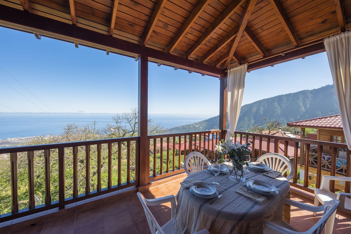 Un área de comedor al aire libre cubierta, con una mesa dispuesta para dos, con vistas panorámicas al mar y a las montañas.