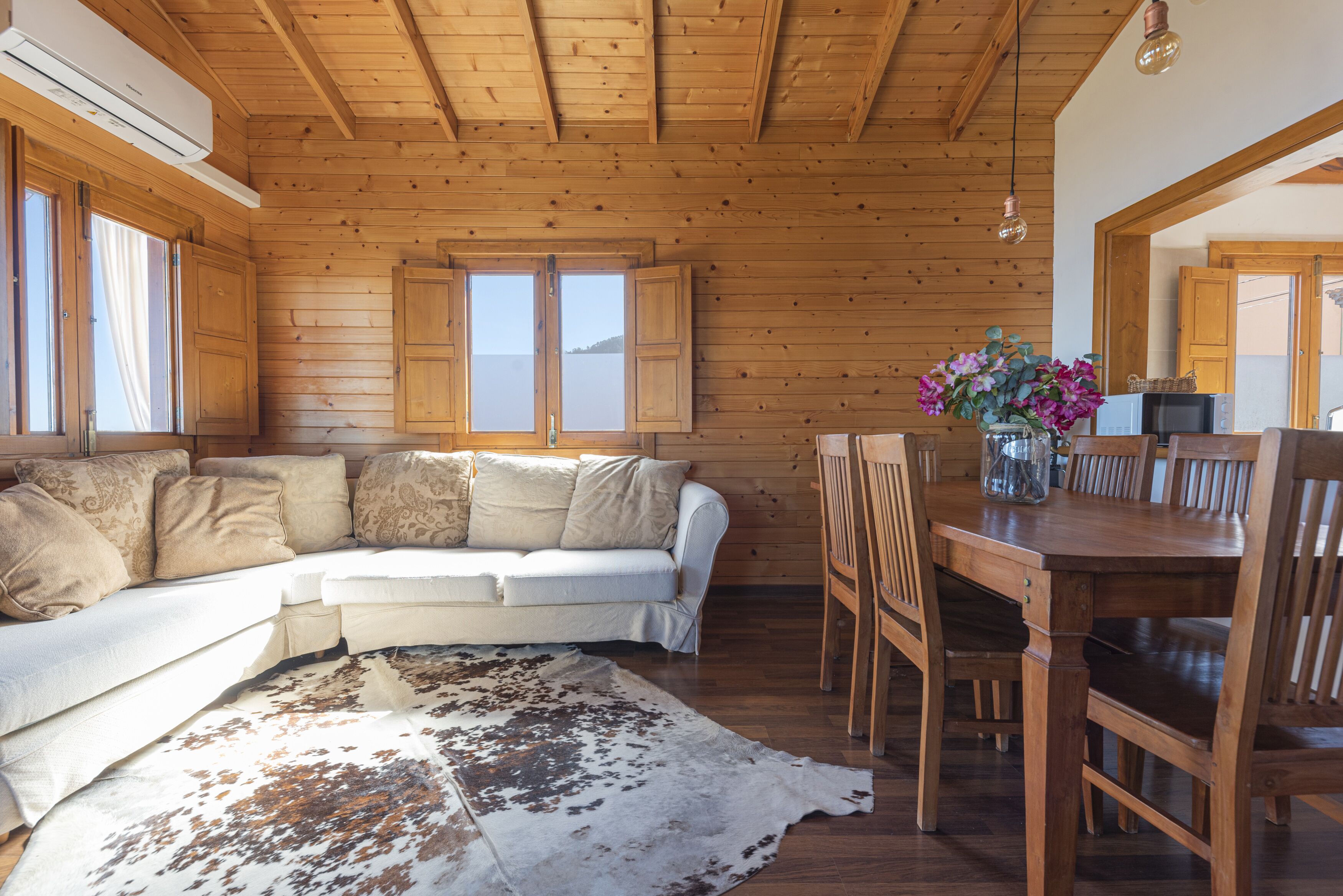 A wooden cabin interior with a sofa, dining table, chairs, and a cowhide rug.