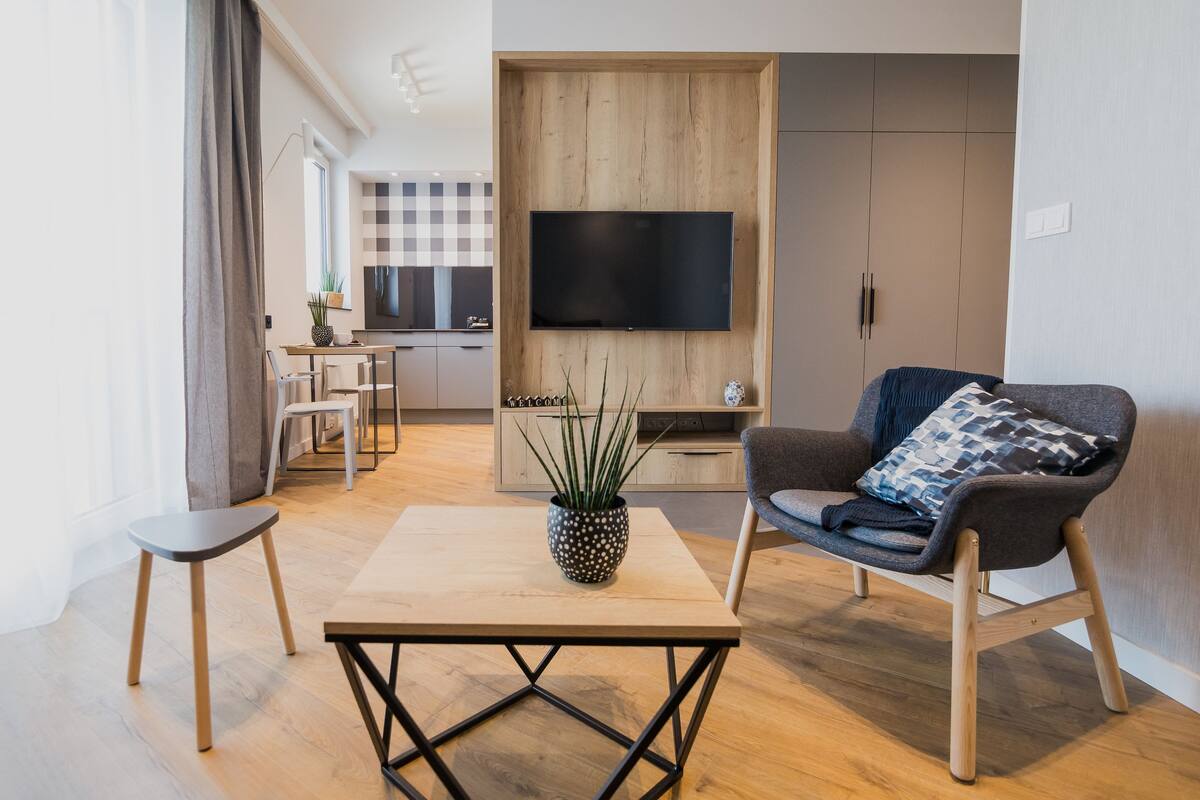 A modern living room with a wooden coffee table, a grey armchair with a patterned throw, and a TV mounted on a wooden wall.
