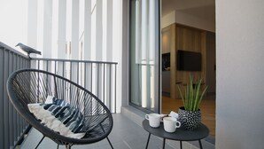 Un balcone con una sedia nera, un tavolino con due tazze bianche e una pentola a pois, e vista su un interno moderno con un televisore.