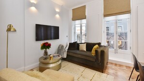 A modern living room with a sofa, a round side table with books and a vase, a flat-screen TV mounted on the wall, and a large window with a view of the outside. - Light-Filled, Contemporary Retreat near The Ritz (London)