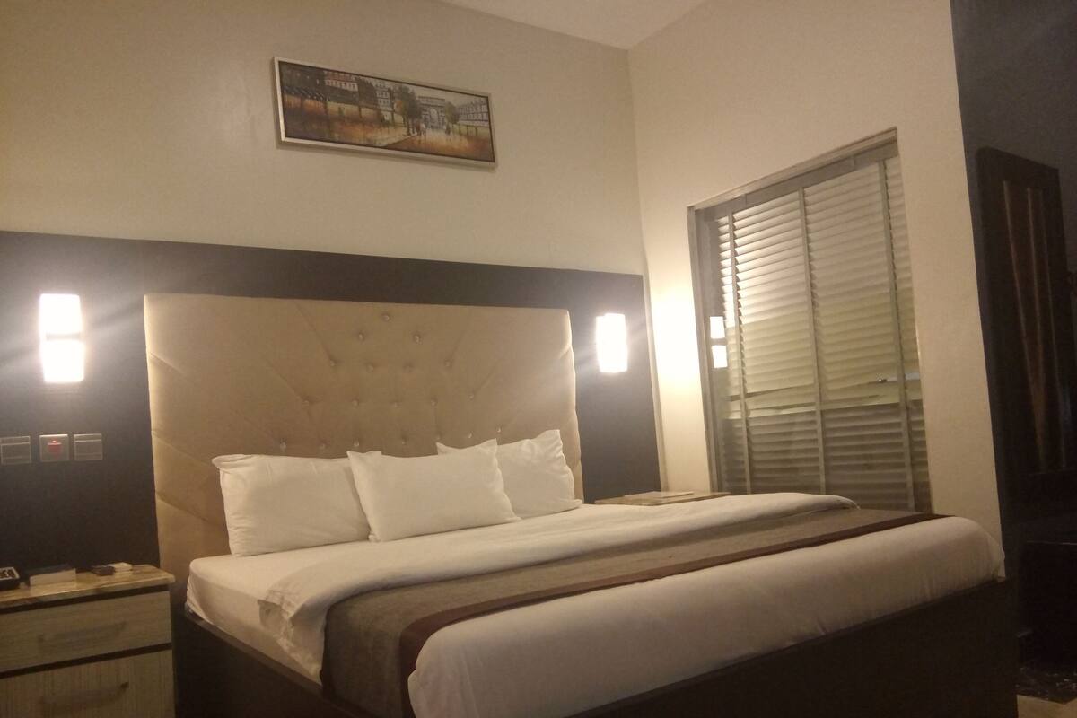 Una habitación de hotel con una cama grande, una mesita de noche con lámpara y una ventana con persianas.