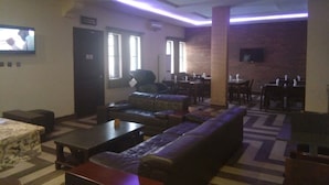 A spacious lounge area with a large sofa, a coffee table, and a television mounted on the wall. There are multiple tables and chairs arranged for dining or socializing. - De Gladys Hotel (Enugu)