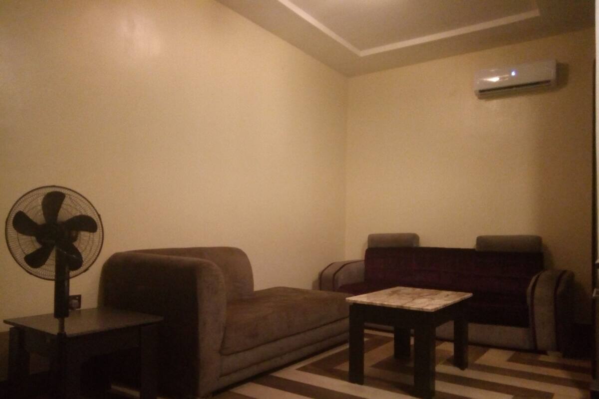 Una sala de estar con un sofá, una mesita, un ventilador y un equipo de aire acondicionado en la pared.