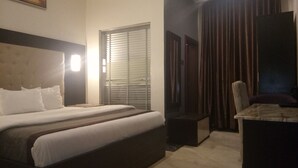 A hotel room with a large bed, a desk, a chair, and a window with blinds. - De Gladys Hotel (Enugu)