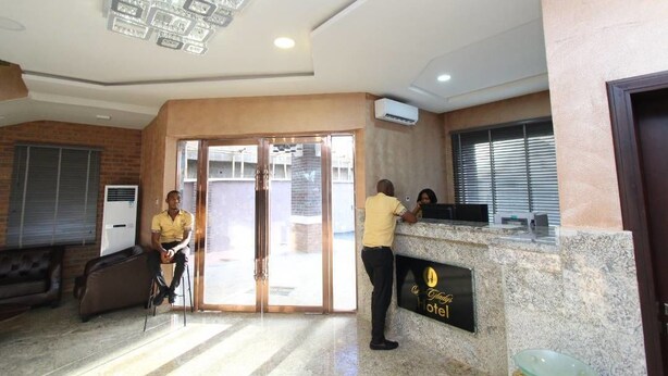 A hotel reception area with a receptionist, a guest seated, and a counter with a sign. - De Gladys Hotel (Enugu)
