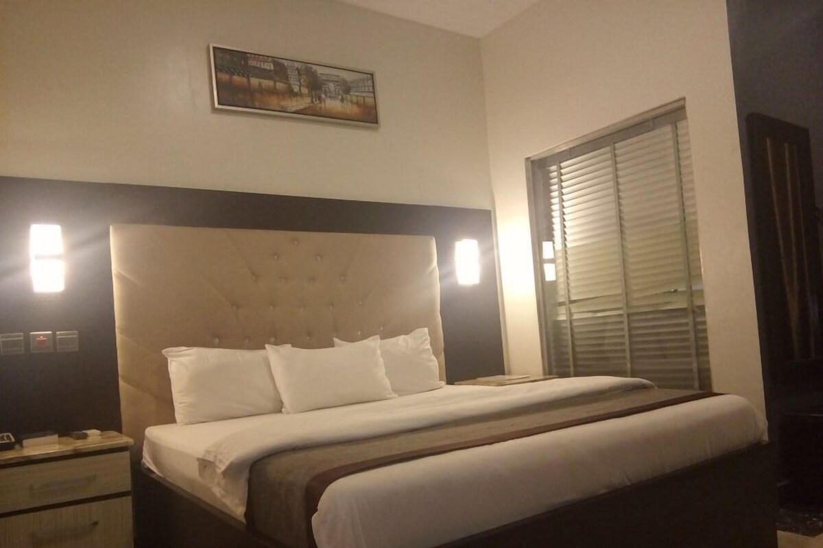 Quarto de hotel com cama grande, mesas de cabeceira, cabeceira, janela com persianas e um quadro pendurado na parede.