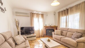 A living room with two beige sofas, a coffee table, a flat-screen TV, a fireplace, and a large window with curtains. - Cubo's Villa Gertrudis. Air conditioning, private pool, wifi. (Alhaurín el Grande)