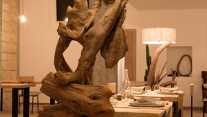 Un comedor con una escultura de madera central, mesas dispuestas con platos y vasos, y una lámpara al fondo.