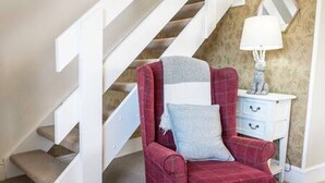 A red armchair with a throw blanket and a pillow, positioned next to a white nightstand with a lamp. A staircase with white railings is visible in the background. - Old Bakery Cottage in Bath - 3 Bedroom, Free (Bath)