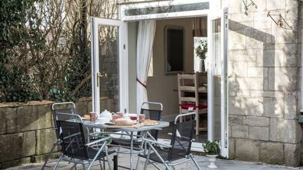 An outdoor patio with a round table and chairs, a glass door leading to a room, and a stone wall. - Old Bakery Cottage in Bath - 3 Bedroom, Free (Bath)