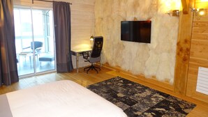 A bedroom with a bed, a desk, a chair, a TV mounted on a stone wall, and a sliding glass door leading to a balcony. - Les Chambres d'Hôtes du Mont Monnet (Longes)