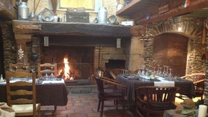 A rustic dining area with a brick floor, wooden tables and chairs, a fireplace, and various kitchen utensils on shelves above. - Les Chambres d'Hôtes du Mont Monnet (Longes)