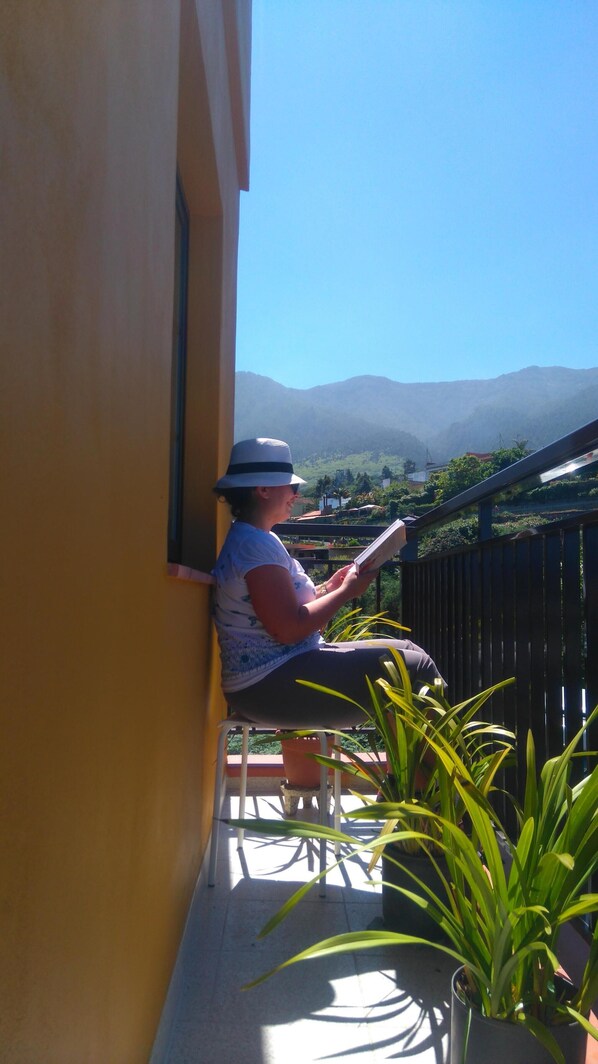 A person sitting on a bench on a balcony with a view of mountains and greenery.
