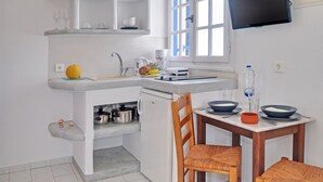 Cocina compacta con horno empotrado, cafetera y un refrigerador pequeño. Hay una mesa de comedor con sillas y un televisor montado en la pared.
