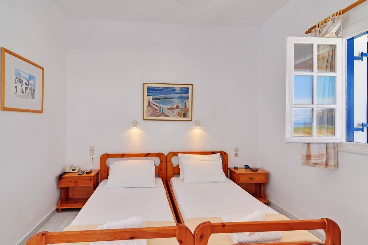 A room with two wooden beds, a nightstand with a phone, a painting on the wall, and a window with curtains.