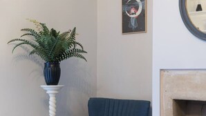 Quarto de canto com uma cadeira azul, uma mesinha redonda branca com uma planta, uma obra de arte emoldurada e uma lareira.