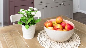 Uma cozinha com mesa de madeira, uma tigela de frutas, uma planta em vaso e um armário.