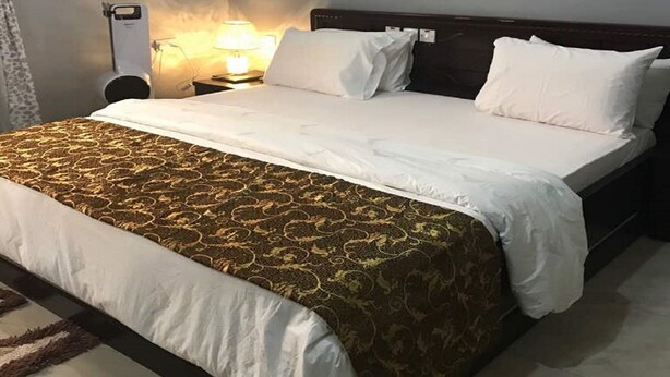 A hotel room with a bed, a nightstand, a lamp, and an air conditioning unit. - Jim-Eddy Suites (Enugu)
