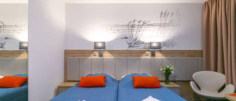 Ein Hotelzimmer mit zwei Betten, jeweils mit blauer Bettwäsche und orangefarbenen Kissen, einem großen Spiegel, einem Wandbild und einem kleinen Tisch mit Blumen.