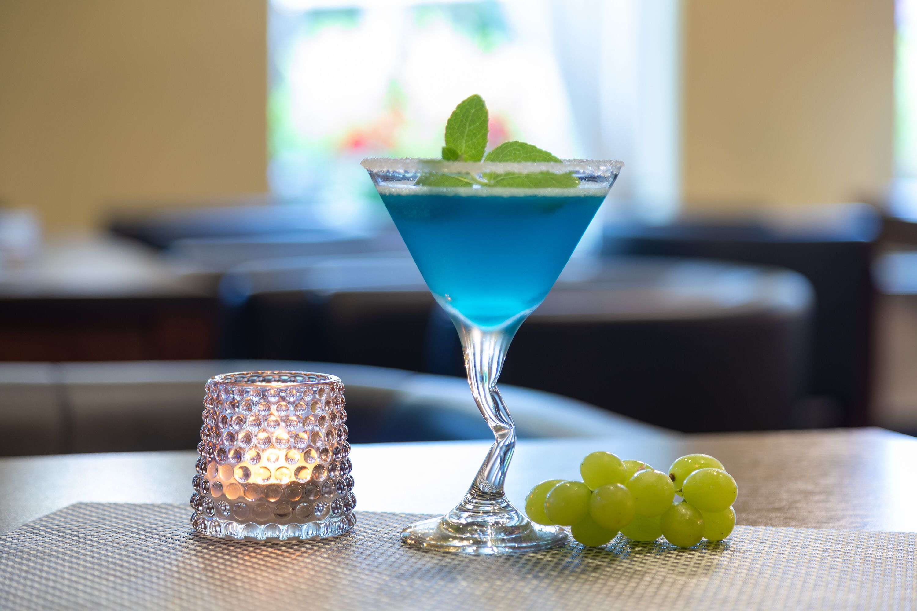 A martini glass with a blue liquid and mint garnish on a table with a textured placemat.
