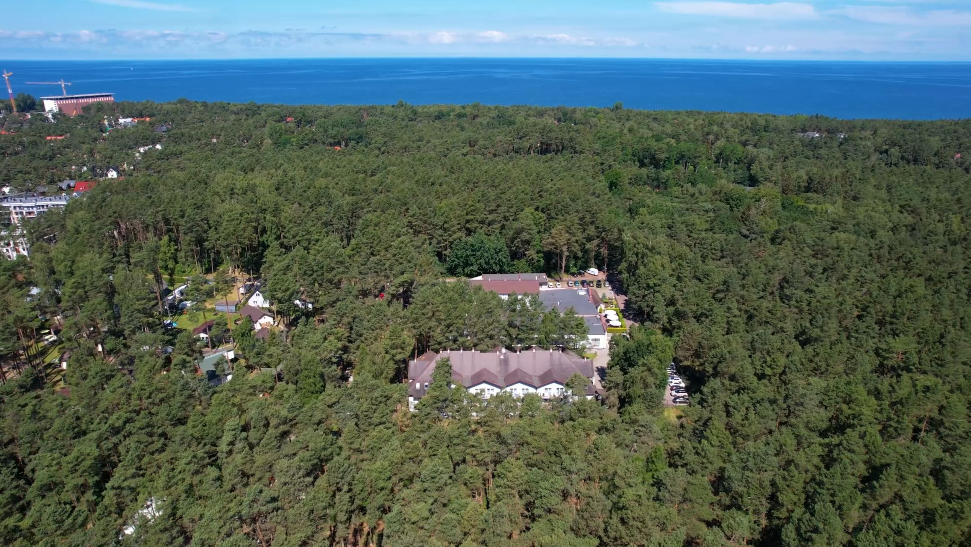 An aerial view of a residential area with dense forestation, a large building with a distinct roof, and a body of water in the background.